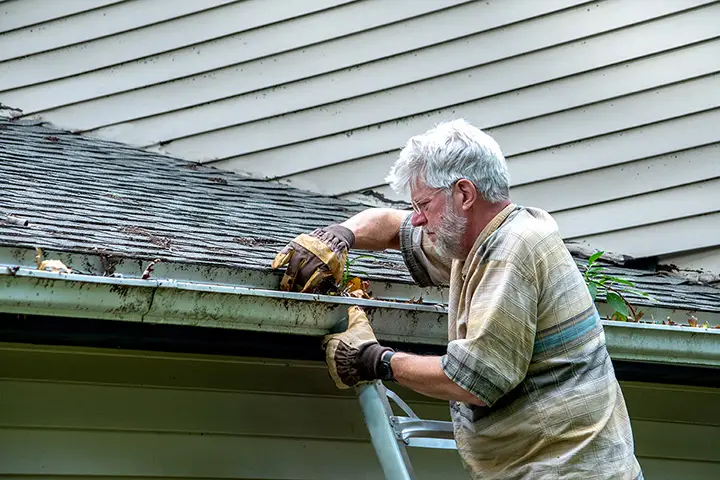 A man stands on a ladder as he cleans out leaves and debris in the gutters on his home